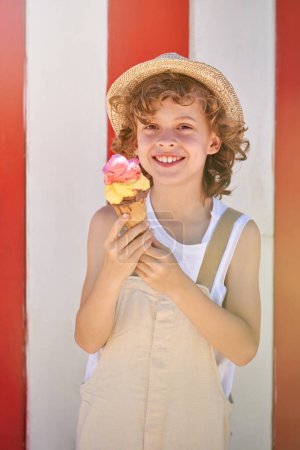 Photo for Happy kid in straw hat smiling and looking at camera while standing against red and white striped background and eating ice cream in waffle cone on summer day - Royalty Free Image