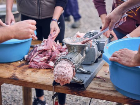 Photo for Crop anonymous coworkers standing around table with pile of beef meat and making mince using metal grinder in yard of butchery house in Mallorca - Royalty Free Image