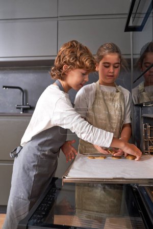 Photo for Focused diverse boy and girl in aprons putting raw cookies on baking sheet of oven while cooking together in kitchen - Royalty Free Image