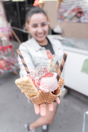 Photo for A young Hispanic woman is smiling holding a traditional ice cream in front of a street market stall. Concept of traditional Mexican dessert - Royalty Free Image