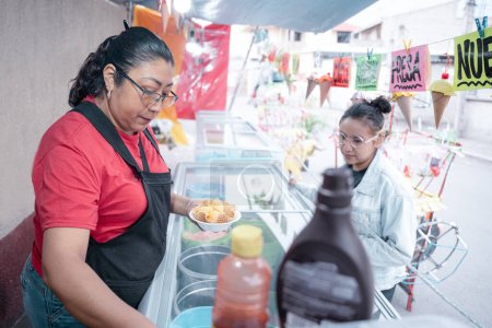 Téléchargez les photos : Une femme hispanique adulte prépare une glace à la vanille pour une jeune cliente dans une échoppe de marché de rue. Concept de dessert mexicain traditionnel - en image libre de droit