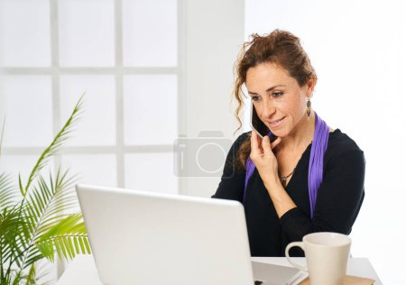 Photo for A woman smiling while answering a phone call and typing on a laptop computer - Royalty Free Image