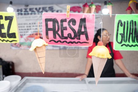 Foto de Carteles de sabor a nuez y fresa en un puesto del mercado de helados con una mujer hispana en el fondo. Concepto de postre tradicional mexicano - Imagen libre de derechos