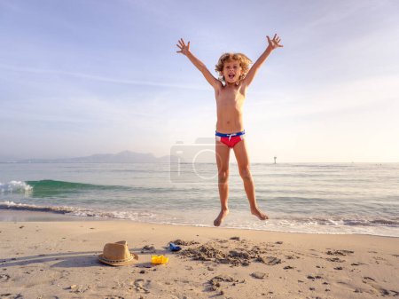 Foto de Cuerpo completo de niño emocionado gritando mientras salta alto en la orilla arenosa húmeda del mar ondulante durante las vacaciones de verano - Imagen libre de derechos