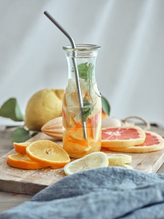 Foto de Vaso de limonada saludable con hojas de naranja limón y menta y frutas en rodajas sobre tabla de cortar de madera - Imagen libre de derechos