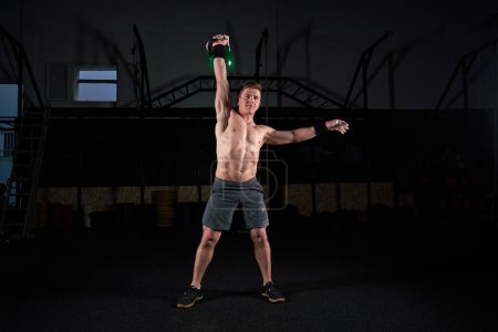 Photo for Muscular athlete with naked torso doing exercise with kettlebell and looking at camera in dark sport club - Royalty Free Image