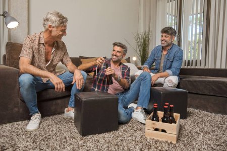 Téléchargez les photos : Corps complet d'hommes riants en vêtements décontractés s'amusant et buvant de la bière tout en passant du temps ensemble dans le salon à la maison - en image libre de droit