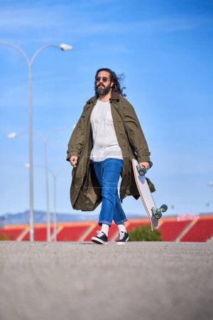 Foto de Hombre barbudo de moda adulto de cuerpo completo en gafas de sol con impermeable elegante y jeans paseando por el camino de asfalto con longboard en un día soleado - Imagen libre de derechos