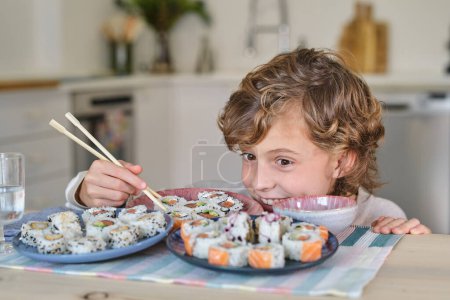 Foto de Chico excitado con el pelo rizado sentado en la mesa de madera y escondido detrás de los platos mientras come deliciosos rollos de sushi con palillos en la cocina en casa - Imagen libre de derechos