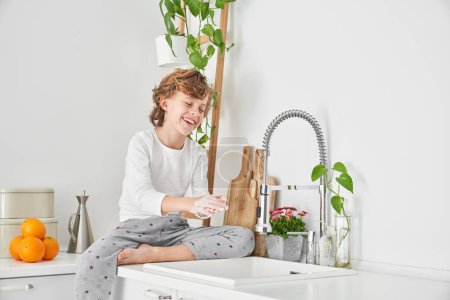 Foto de Niño descalzo de pelo rizado sentado cerca del lavabo y lavándose las manos con jabón para prevenir la propagación de bacterias en la cocina por la mañana - Imagen libre de derechos