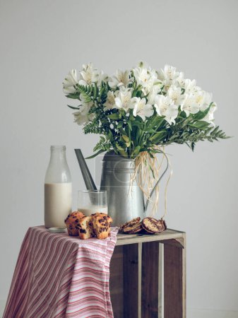 Foto de Composición de flores blancas en regadera colocada cerca de botella de leche y magdalenas en caja rústica sobre fondo blanco - Imagen libre de derechos