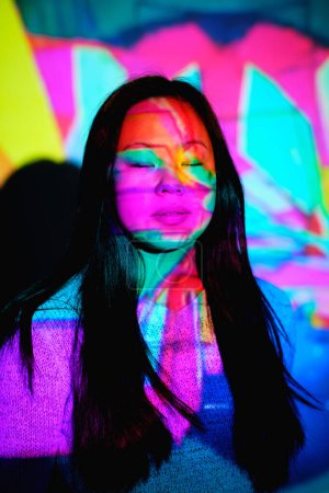 Téléchargez les photos : Tranquille femelle asiatique avec de longs cheveux noirs debout avec les yeux fermés près du mur sous un éclairage au néon multicolore lumineux dans une chambre élégante - en image libre de droit