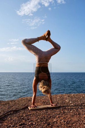 Téléchargez les photos : Vue arrière d'une athlète féminine sans visage en tenue de sport debout sur une planche de bois à Adho Mukha Vrikshasana posant avec des jambes à Baddha Konasana tout en pratiquant le yoga sur la plage - en image libre de droit