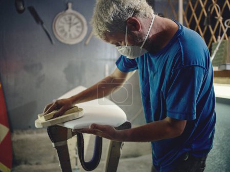 Foto de Vista lateral del trabajador del hombre en la máscara respiratoria protectora pulido pieza de trabajo de tabla de surf con abrasivo en el taller - Imagen libre de derechos