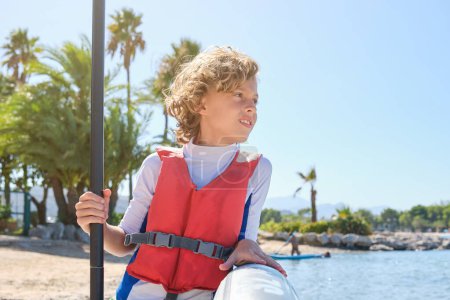 Téléchargez les photos : Preteen enfant avec planche à pagaie portant gilet de sauvetage regardant loin tout en se tenant debout sur le bord de mer sablonneux le jour ensoleillé d'été - en image libre de droit