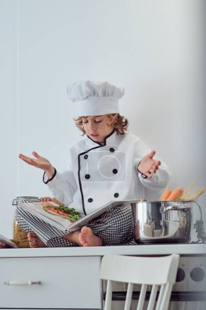 Foto de Desde abajo de cuerpo completo niño preadolescente en traje de chef y sombrero sentado en el mostrador de la cocina y la lectura de libro culinario y expresar desconcierto de la receta - Imagen libre de derechos