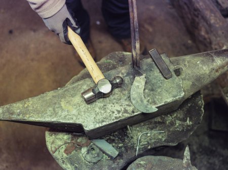 Photo for From above of crop anonymous blacksmith in protective gloves using hammer and iron sickle while working on anvil in workshop - Royalty Free Image