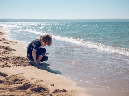 Foto de Vista lateral cuerpo completo de niño encantador sentado en el suelo y jugando con arena mojada cerca del mar ondulante en el día de verano soleado - Imagen libre de derechos