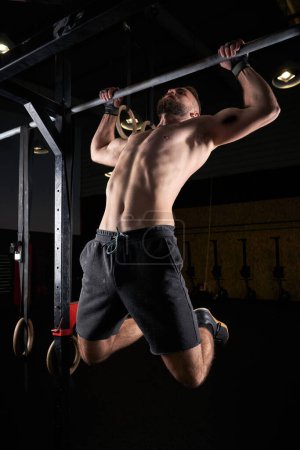 Foto de Cuerpo completo de atleta masculino musculoso sin camisa haciendo flexiones en la barra durante el entrenamiento intenso en el gimnasio oscuro - Imagen libre de derechos