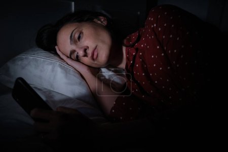 Téléchargez les photos : Sérieux hispanique femme surf internet sur téléphone portable moderne sur le lit tard dans la nuit tout en souffrant d'insomnie dans la chambre sombre - en image libre de droit
