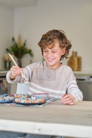 Foto de Niño feliz con ropa casual sonriendo mientras está sentado en la mesa de madera y comiendo deliciosos rollos de sushi con palillos en el comedor en casa - Imagen libre de derechos