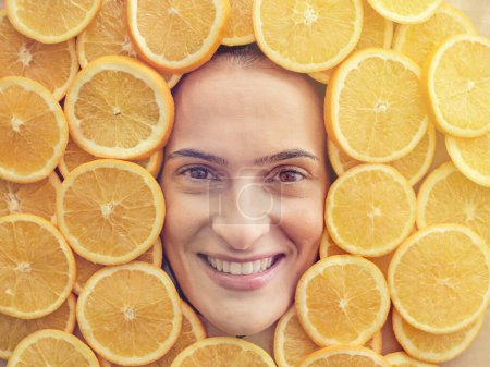 Foto de Rodajas de naranjas jugosas sanas maduras colocadas alrededor de la cara del modelo femenino feliz mirando a la cámara con una amplia sonrisa dentada - Imagen libre de derechos