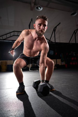 Photo for Full body shirtless bearded sportsman preparing to lift hexagon dumbbell during functional workout in dim light of fitness center - Royalty Free Image