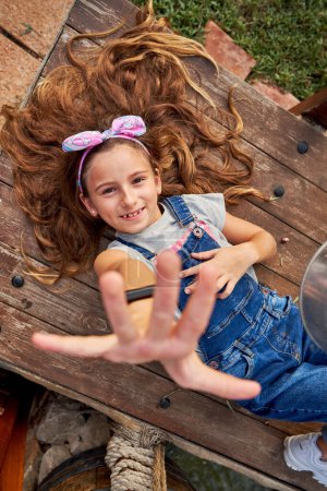 Photo for Top view of cheerful girl in denim overall looking at camera with raised arm while resting on wooden bench in countryside - Royalty Free Image