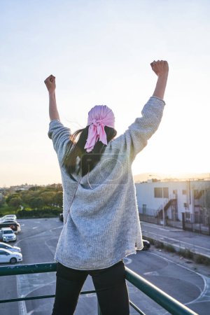 Photo for Back view of anonymous female achieved remission of breast cancer wearing pink scarf raising fists up while standing on balcony against parking lot at sundown - Royalty Free Image