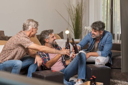 Téléchargez les photos : Hommes d'âge moyen s'amuser ensemble tout en buvant de la bière et se détendre dans le salon à la maison - en image libre de droit