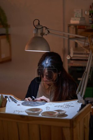Foto de Joyero femenino étnico concentrado creando patrón en accesorio mientras usa taladro giratorio durante el trabajo en la mesa de madera en el estudio - Imagen libre de derechos