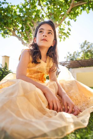 Foto de Bajo ángulo de niño de ensueño en vestido amarillo sentado en el césped cubierto de hierba y mirando hacia otro lado en el patio trasero verde en el día de verano brillante - Imagen libre de derechos