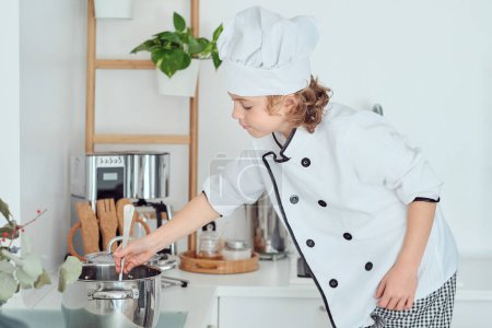 Foto de Vista lateral de un niño pequeño en uniforme de chef blanco revolviendo la comida en la sartén con cuchara mientras se cocina en la cocina en casa - Imagen libre de derechos