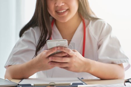 Photo for Crop unrecognizable positive female medic in white uniform texting message on cellphone at table with clipboard during work in light clinic - Royalty Free Image