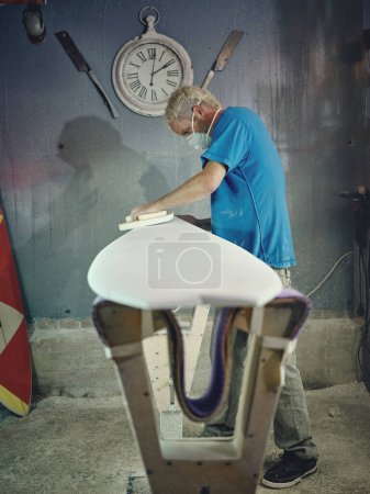 Photo for Side view of focused young male master with blond hair in t shirt and respirator sanding edges of surfboard while working in traditional studio - Royalty Free Image