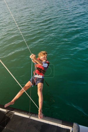 Photo for From above of active preteen boy with blond hair in life vest hanging on rope from yacht over wavy sea and looking away on sunny day - Royalty Free Image