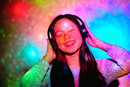 Téléchargez les photos : Optimiste asiatique femme écoute chanson dans les écouteurs avec les yeux fermés tout en se tenant près du mur en studio avec éclairage au néon multicolore - en image libre de droit