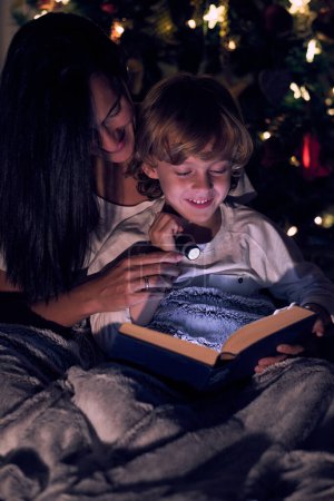 Téléchargez les photos : Mère positive et fils mignon lisant livre intéressant avec lampe de poche tout en étant assis sous la couverture près de l'arbre de Noël avec guirlande lumineuse - en image libre de droit
