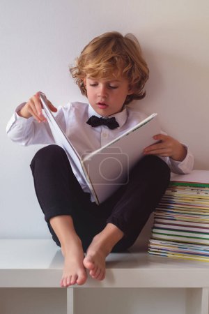 Foto de Cuerpo completo de adorable chico descalzo en ropa formal leyendo interesante libro mientras está sentado sobre fondo blanco en la sala de luz - Imagen libre de derechos