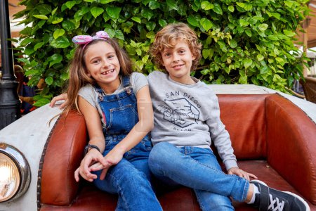 Photo for Happy cute friendly kids with legs crossed in casual clothes smiling and looking at camera while sitting on retro sofa in shape of car - Royalty Free Image