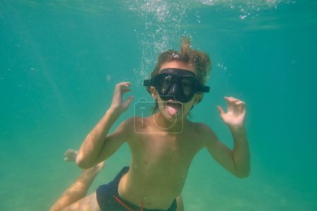 Photo for Excited child in swimwear and diving mask swimming with tongue out in blue ocean during summer vacation while looking at camera - Royalty Free Image