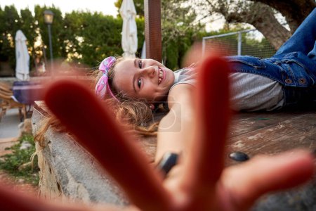 Téléchargez les photos : Préadolescent souriant en denim regardant globalement la caméra et tendre la main floue tout en étant allongé sur un banc en bois à la campagne - en image libre de droit