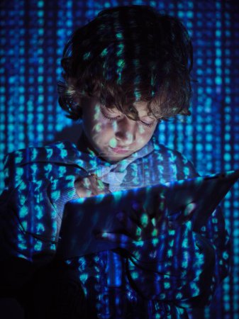 Téléchargez les photos : Jeune programmeur garçon en sweat à capuche en utilisant une tablette numérique pour le codage de logiciels malveillants dans la projection de symboles informatiques dans la chambre noire - en image libre de droit