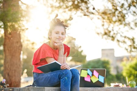 Téléchargez les photos : Souriant fille intelligente assis sur un banc en bois avec livre ouvert près d'un ordinateur portable avec des notes collantes tout en faisant des devoirs dans le parc le jour ensoleillé - en image libre de droit