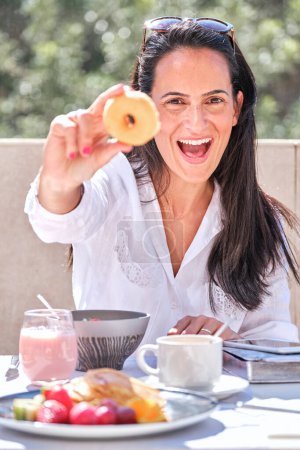 Foto de Feliz hembra desayunando con yogur y frutas y bayas mostrando sabrosa dona y mirando a la cámara mientras está sentado en la mesa en la terraza del hotel en el día soleado - Imagen libre de derechos