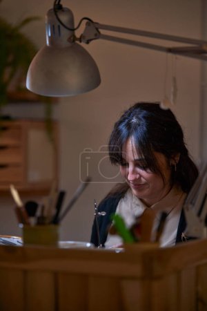 Photo for Professional female master sitting at table with instrument under lamp light and creating handmade jewelry accessories - Royalty Free Image