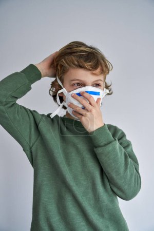 Téléchargez les photos : Enfant en vêtements décontractés portant un respirateur médical pendant les mesures restrictives de verrouillage et faisant face à une nouvelle normale - en image libre de droit