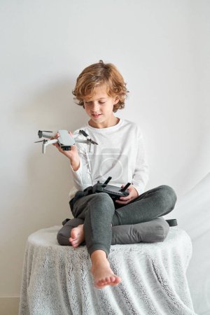 Foto de Cuerpo completo descalzo chico sentado en la mesa contra la pared blanca y mirando hacia abajo drone juguete en la mano en la sala de luz - Imagen libre de derechos