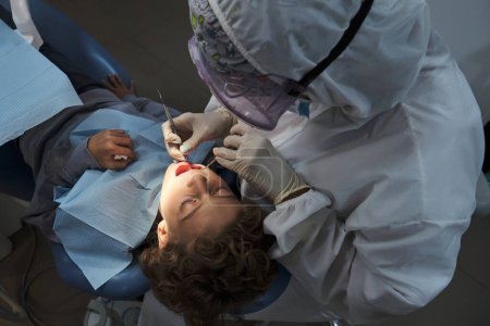 Foto de De arriba del dentista anónimo en el uniforme y los guantes usando la herramienta dental para comprobar los dientes del muchacho en la clínica - Imagen libre de derechos