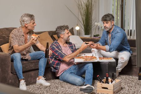 Téléchargez les photos : Heureux hommes d'âge moyen et en vêtements décontractés manger de délicieuses pizzas tout en passant du temps ensemble à la maison - en image libre de droit